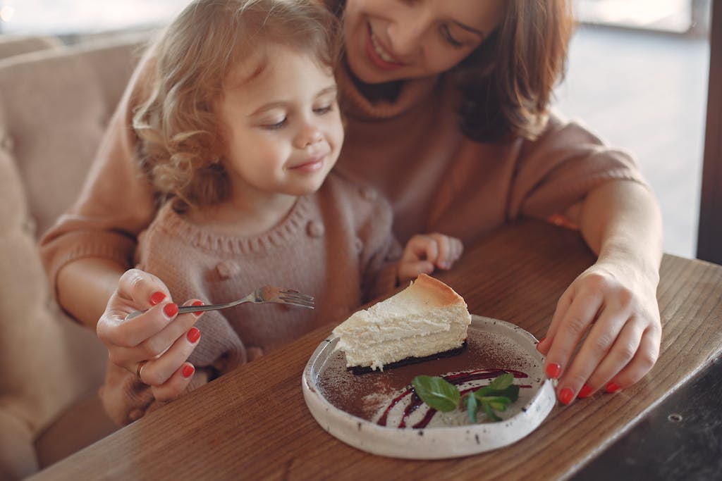 Crop happy mother in casual clothes sitting at table and eating piece of cake with little girl while spending time at cafeteria together